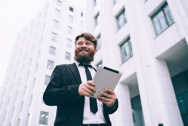 From below of cheerful bearded man with brown hair in formal wear walking in downtown near buildings and using tablet - Foto, imagen