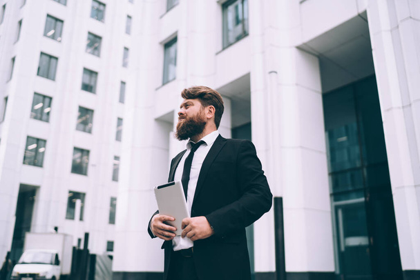 Low angle of serious plump unshaven male entrepreneur in office suit with tablet in hands standing near facade of modern city building and looking away - Фото, изображение