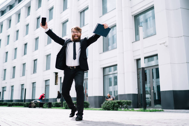 Low angle of full length happy bearded businessman in formal outfit and eyewear near modern building and celebrating new project with smartphone and notebook in hands - Photo, Image