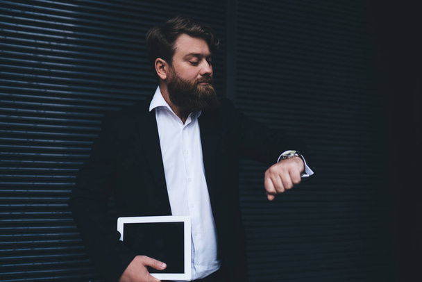 Serious bearded businessman in formal wear with tablet in hand looking at wristwatch while standing against black wall during break - Foto, afbeelding