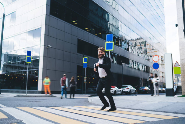 Full body of happy successful businessman with tablet running on pedestrian crossing against modern tower with mirrored walls on city street - Фото, изображение