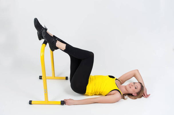 Blond žena dělá cvičení s vysokou žlutou podlahové mříže na bílém pozadí. žena v černých kamaších a žlutém tričku - Fotografie, Obrázek