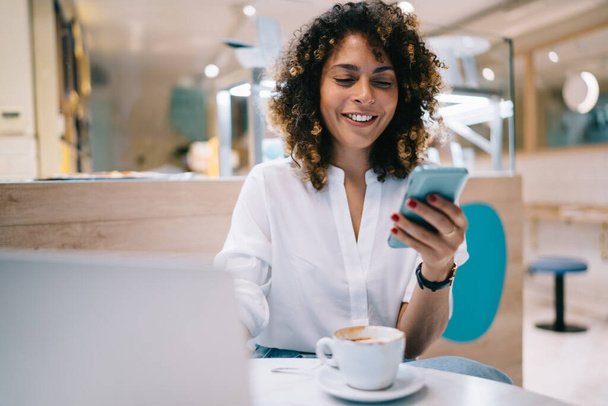Smiling young ethnic female freelancer in casual clothes browsing smartphone while sitting at table in modern cafe near laptop with cup of hot coffee - Φωτογραφία, εικόνα
