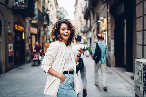 Side view of young stylish ethnic lady in casual clothes standing with smartphone on narrow pedestrian street of old city and looking at camera - Φωτογραφία, εικόνα