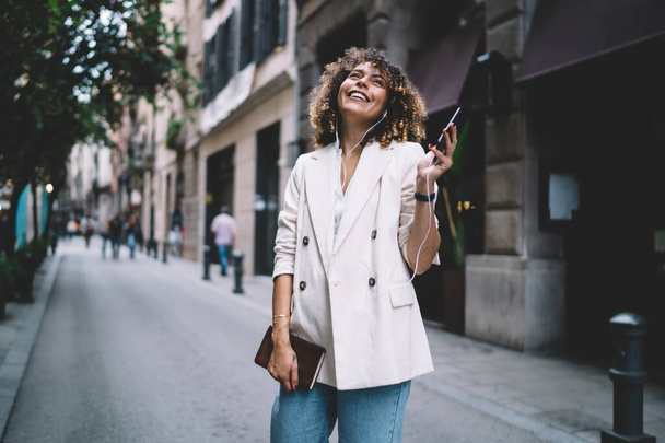 Young female with curly brown hair in stylish white jacket standing on street in old town and smiling while talking on smartphone with earphones - Photo, Image