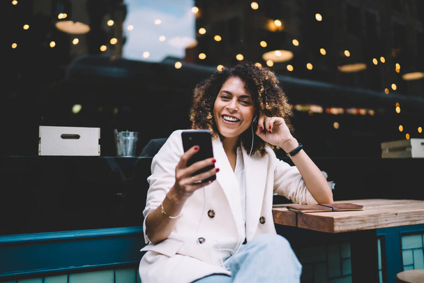 Young woman with earphones in casual outfit and wristwatch browsing smartphone and smiling while sitting at table near glass wall of cafe on street - Photo, Image