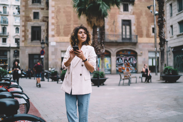 Young female tourist in casual clothes looking away and using smartphone while traveling on square with palms in old city - Foto, Imagen