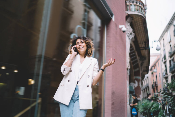 Low angle view of cheerful ethnic young lady in stylish outfit looking away and emotionally talking on phone while walking along street in city - Фото, изображение