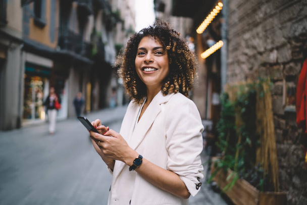 Young ethnic woman with smartphone smiling and standing on street and browsing internet wearing casual clothes on sunny day looking forward - Foto, Imagen