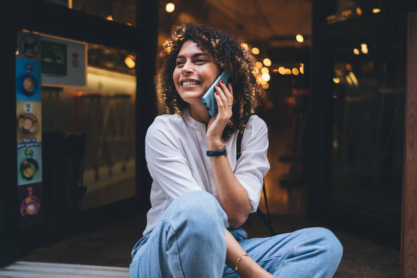 Positive young ethnic woman wearing casual talking with friend on mobile phone and smiling sitting on floor outside near creative cafe - Foto, Bild