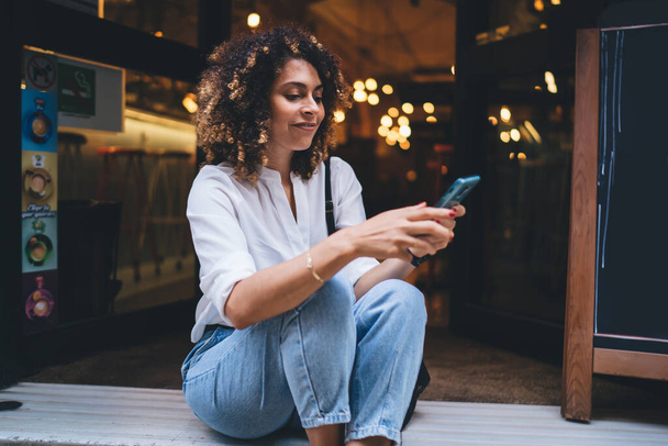 Cheerful positive ethnic woman wearing casual clothes sitting on floor and browsing smartphone while resting in cafe during weekend at evening - Fotoğraf, Görsel