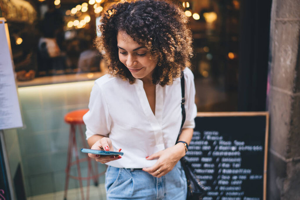 Позитивна етнічна жінка зі смартфоном посміхається і переглядає Інтернет під час стояння на вулиці біля кафе в повсякденному одязі, дивлячись на екран
 - Фото, зображення