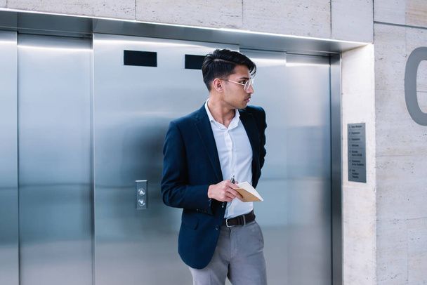 Serious ethnic male entrepreneur in formal clothing and eyeglasses standing near elevator in modern office and holding notebook in hands - Foto, Imagen