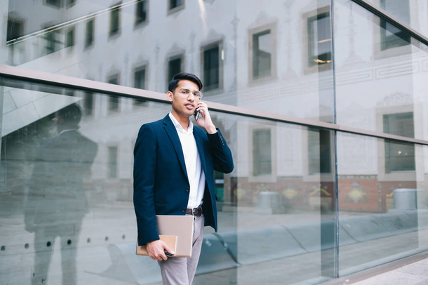 Half length portrait of handsome 20s hindy male proud ceo in trendy outfit standing outdoors talking on mobile phone, positive businessman looking at camera making smartphone conversation in roaming - Fotó, kép