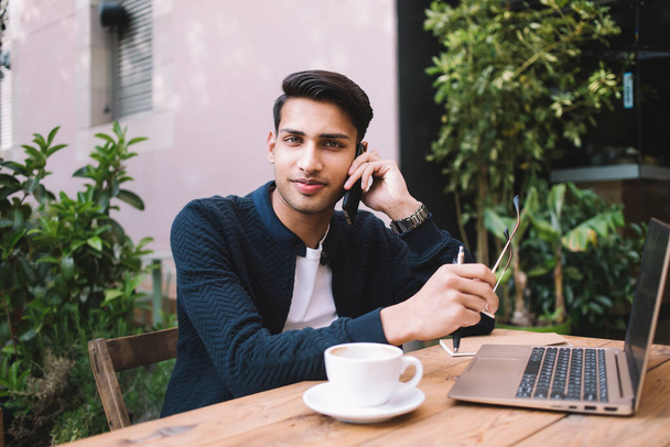 Portrait of positive hindu male millennial blogger working remotely on laptop computer on cafe terrace making smartphone call, smiling businessman talking on smartphone on distance job process - Zdjęcie, obraz