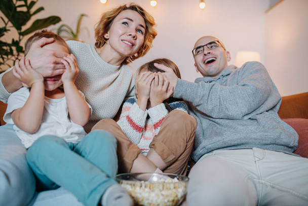 Unexpected moment while family watching television together on the couch in the living room. Laughing parents covering faces of their two children. - Fotografie, Obrázek