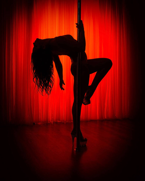 Pole dance. Silhouette of a slender sexy woman dancing on a pole in the interior of a nightclub with a red background. - Foto, Bild