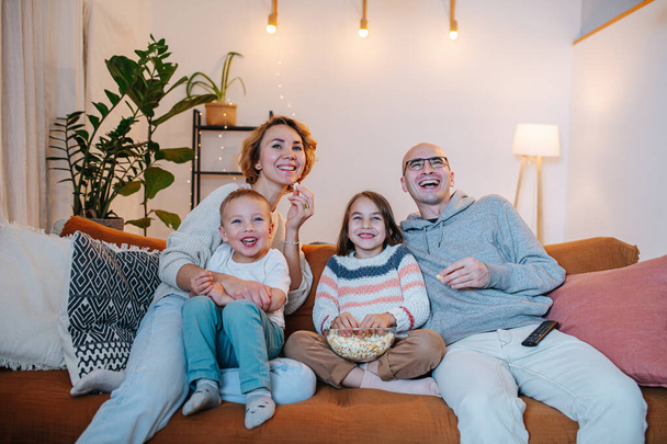 Amused family of parents and two children watching comedy together on the couch in the living room. - Valokuva, kuva