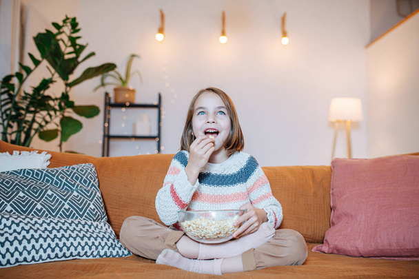 Staring entertained little girl watching TV and eating popcorn, sitting on a couch in a living room - Fotografie, Obrázek