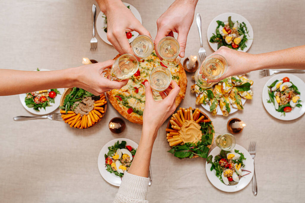 Overhead view of hands clinking glasses over a table with Italian cuisine assorti. Margarita, cheese sticks with hummus and arugula, salad with tomatoes and eggs served with white wine. - Foto, afbeelding
