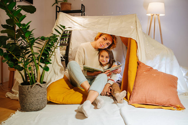 Portrait of mother and daughter sitting in a tent made of sheets with a book at home, posing for a photo, looking at camera. Dim light and garland atop. - Fotoğraf, Görsel