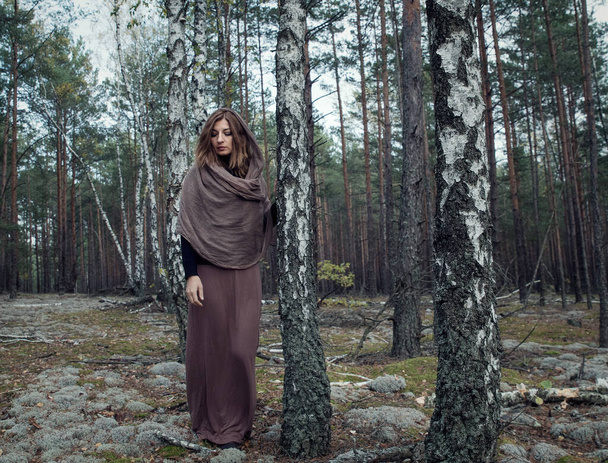 Une fille en robe longue avec une écharpe brune sur la tête dans une sombre forêt de bouleaux d'automne. - Photo, image