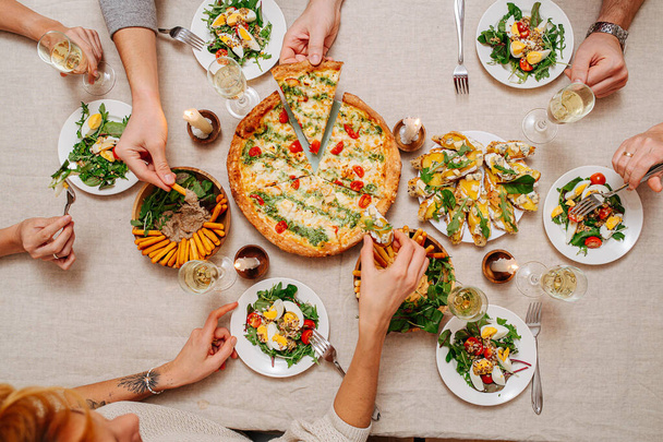 Overhead view of hands of dining people over a table with Italian cuisine assorti. They are eaing margarita, cheese sticks with hummus and arugula, salad with tomatoes and eggs served with white wine. - Zdjęcie, obraz