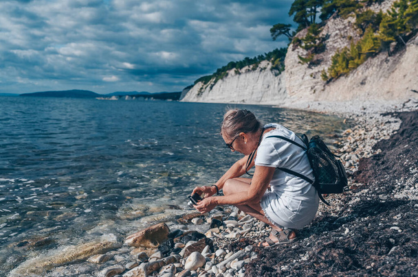 Anciana mayor viajando mochilero mujer madura turista tomando fotos costa del mar, rocas, cielo azul - Foto, Imagen