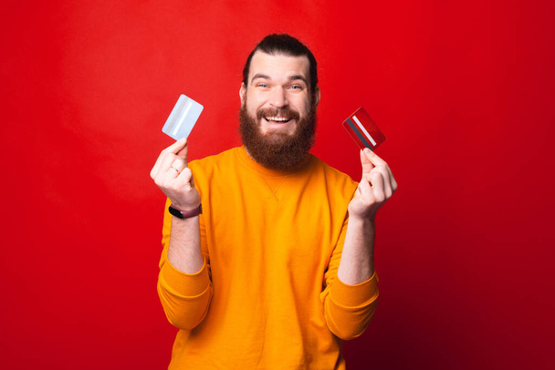 Podekscytowany młody brodaty mężczyzna patrzy w kamerę i trzyma dwie karty kredytowe - Zdjęcie, obraz