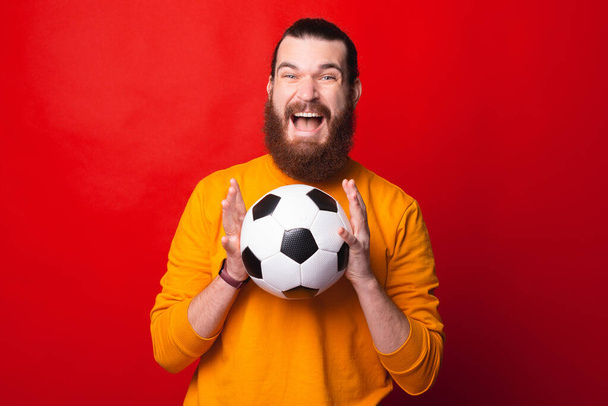 Un hombre emocionado está mirando a la cámara sosteniendo una pelota de fútbol cerca de una pared roja - Foto, imagen