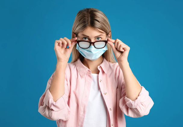 Woman with foggy glasses caused by wearing disposable mask on blue background. Protective measure during coronavirus pandemic - Фото, изображение