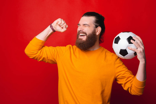 Ένας γενειοφόρος νεαρός άνδρας κρατά μια μπάλα ποδοσφαίρου και είναι ενθουσιασμένος κοντά σε ένα κόκκινο τοίχο - Φωτογραφία, εικόνα