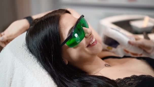 Kobieta kosmetyczka robi laserową depilację pachy atrakcyjnej seksownej kobiety leżącej na medycznej kanapie i uśmiecha się w salonie piękności. Kosmitologia, depilacja i koncepcja spa. Opieka ciała. Zwolnij trochę. Zbliżenie - Materiał filmowy, wideo