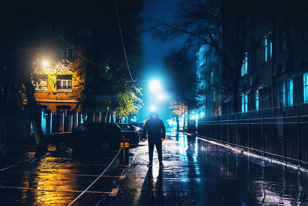 Silhouette of alone stranger in hood at night city street in rain. Creepy killer or stalker, criminal stands in shadow with urban lights reflected in puddles. Thriller horror mysterious atmosphere - Photo, Image