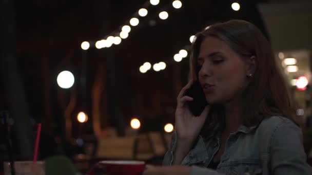 Valkoihoinen nainen puhuu älypuhelimellaan istuessaan jalkakäytävällä kahvilassa kesäiltana. Media. Nuori nainen, jolla on punainen kuppi cappuccinoa tai teetä, on matkapuhelinkeskustelu. - Materiaali, video