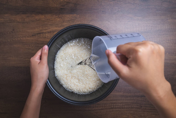 La mano de la mujer está vertiendo agua usando vidrio medidor para cocinar arroz de jazmín. sobre mesa de madera. - Foto, Imagen