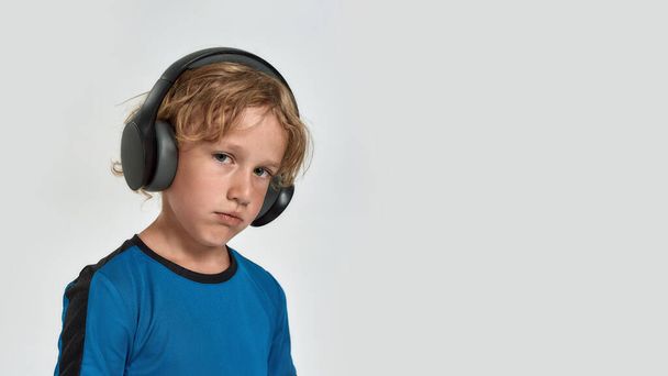 Portrait of tired little sportive boy child in sportswear and headphones looking at camera while posing, standing isolated over white background - 写真・画像