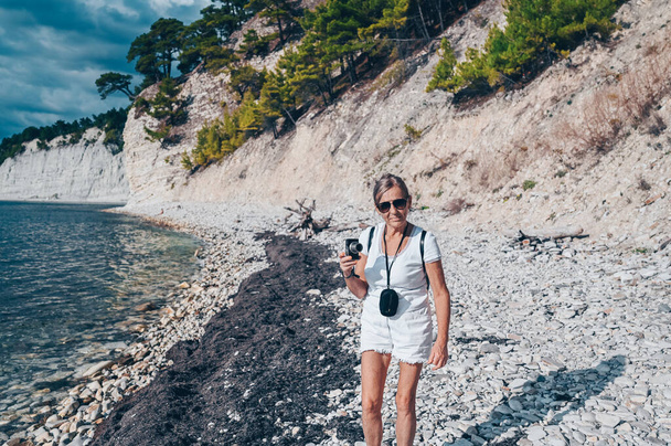 高齢者のシニア旅行バックパッカー成熟した女性の観光客の写真を撮る海の海岸、岩、青空 - 写真・画像