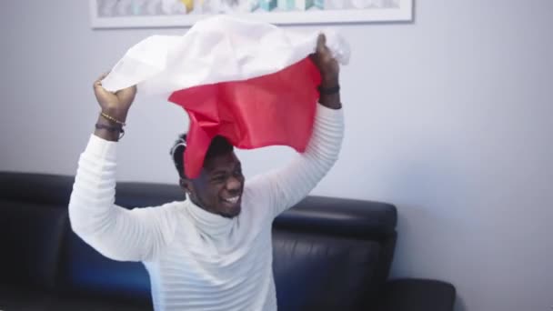 Νεαρός ευτυχισμένος μαύρος άνδρας με πολωνική σημαία βλέποντας ποδόσφαιρο παιχνίδι στην τηλεόραση - Πλάνα, βίντεο