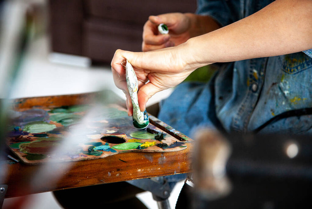 Woman artist hand squeezing a tube with yellow orange oil paint on wooden palette for painting in art studio. Concept of creating new colours by mixing oil paint. Unfocused paintbrushes. - Photo, Image