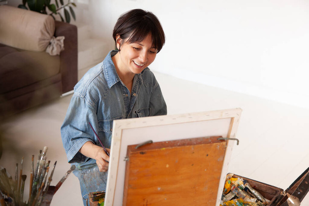 Happy female artist holding framed canvas, paintbrush and ready for painting at home during weekend. Hobby and leisure activity. Excited and inspired painter woman by painting. - Фото, зображення