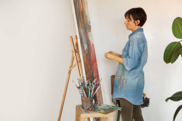 Young woman artist holding paintbrush and painting with oil paint on framed cotton canvas on easel at home. Hobby and leisure concept. Painter creating art. Interior. Copy space. - Photo, image