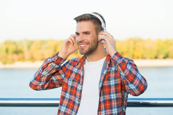 sonriente hombre barbudo escuchar música en auriculares digitales con camisa a cuadros al aire libre, tecnología - Foto, imagen