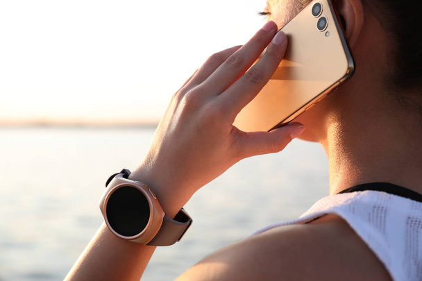 Woman with fitness tracker talking on phone near river, closeup - Foto, imagen
