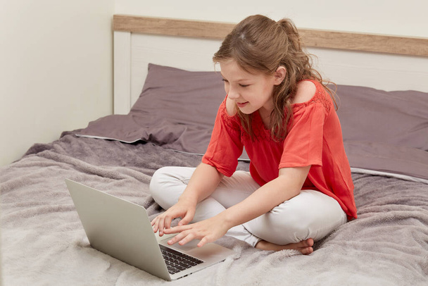 A beautiful girl sits on a light bed in the bedroom and communicates online. - Фото, изображение