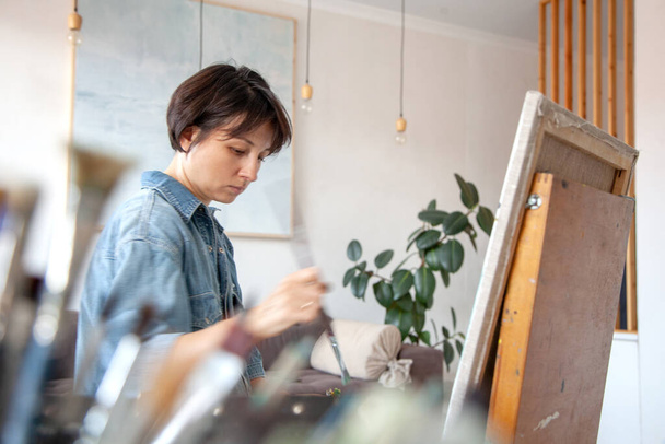 Concentrated short-haired female artist painting on cotton canvas with oil paint at home. Painter - 写真・画像