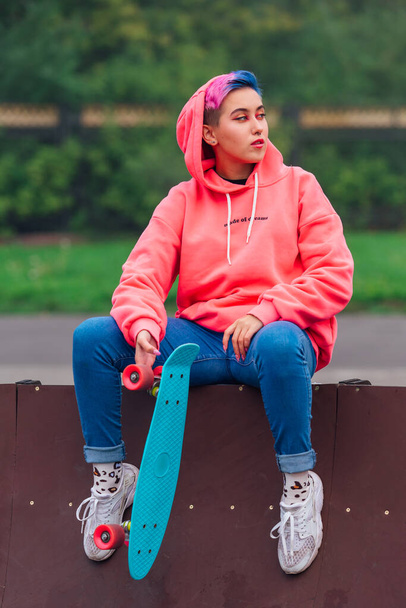 Portrait of a trendy pretty young girl sitting next to the skateboard court with her plastic skateboard. - Foto, Bild