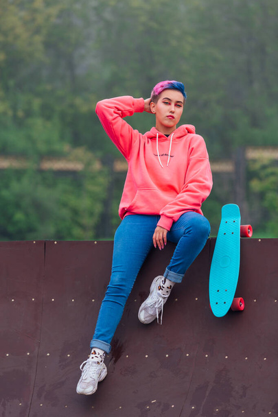 Portrait of a trendy pretty young gir with short colored hair sitting next to the skateboard court with her plastic skateboard. - Φωτογραφία, εικόνα