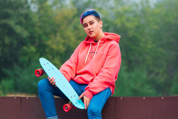 Summer lifestyle image of trendy pretty young girl with short colored hair sitting next to the skateboard court with her plastic skateboard. - Photo, Image