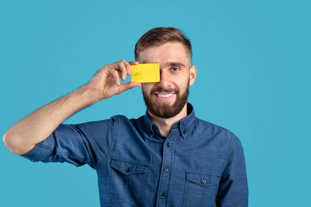 Sorrindo millennial cara segurando cartão de crédito na frente de seu olho no fundo do estúdio azul - Foto, Imagem
